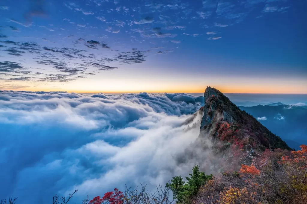 Hallando Fortaleza en la Fragilidad: La Resiliencia Según la Biblia Amanecer majestuoso sobre un mar de nubes, con una montaña escarpada emergiendo como símbolo de resiliencia. El pico rocoso se eleva por encima de la niebla, rodeado de árboles otoñales en rojo y verde. El cielo azul se funde con tonos cálidos del sol naciente, creando un paisaje impresionante que evoca fuerza y perseverancia ante los desafíos de la naturaleza.